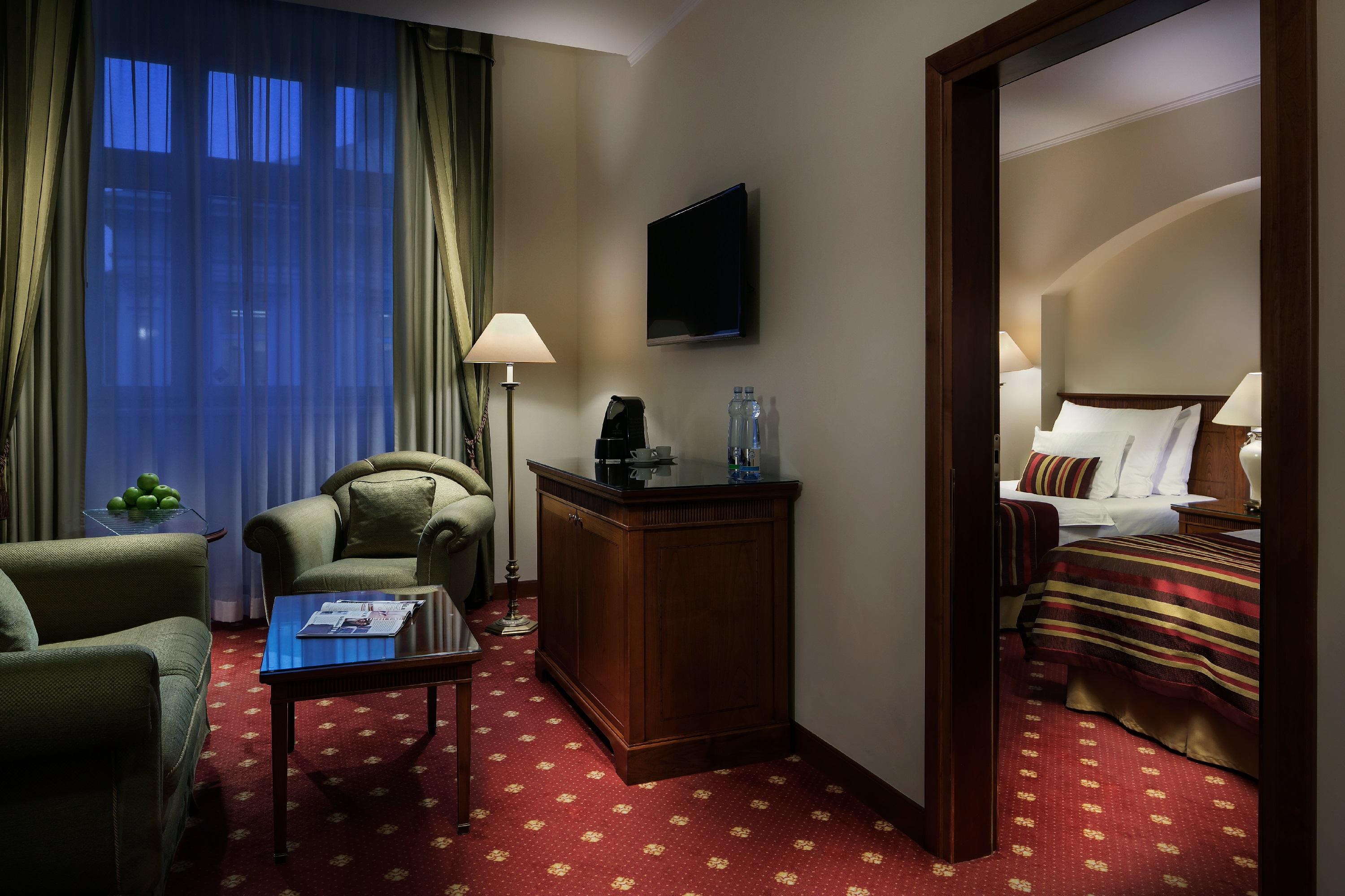 Art Nouveau Palace Hotel Praga Esterno foto The photo shows a room that appears to be part of a hotel suite. On the left side, there is a small seating area with a green armchair and a small table. A lamp stands beside the chair, providing light. In the center, there's a wooden cabinet or dres