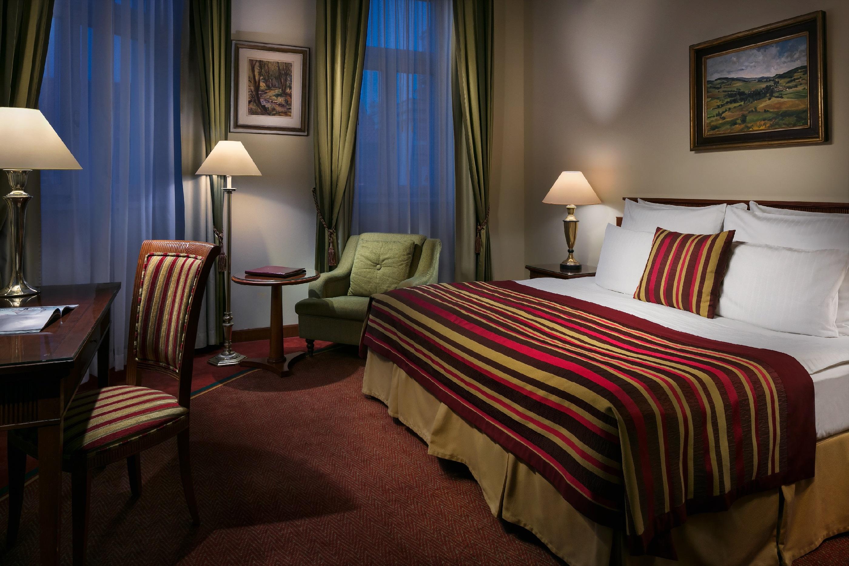 Art Nouveau Palace Hotel Praga Esterno foto The photo shows a cozy hotel room. It features a bed with a colorful striped blanket. There are two lamps on either side of the bed, providing soft lighting. A comfortable green armchair is positioned nearby, along with a small side table. A wooden d