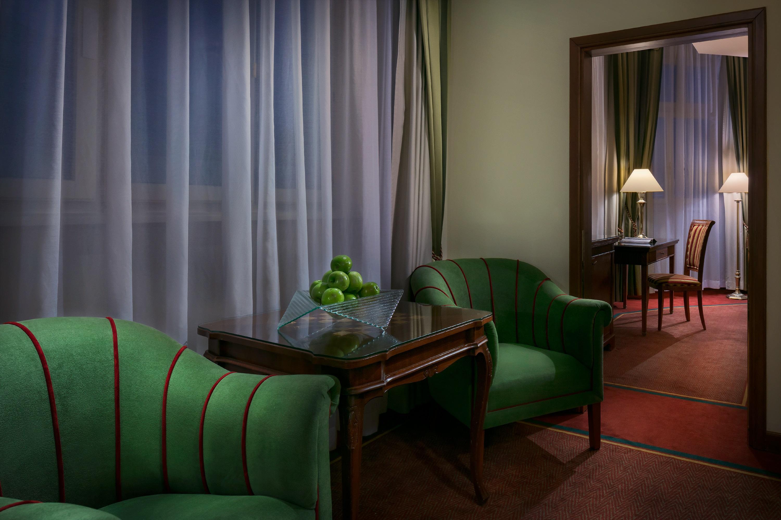 Art Nouveau Palace Hotel Praga Esterno foto The photo depicts a cozy interior space with a warm ambiance. There are two green chairs with red stripes positioned beside a wooden table that holds a bowl of green apples. The room features soft, sheer curtains that create a flowing effect over a w