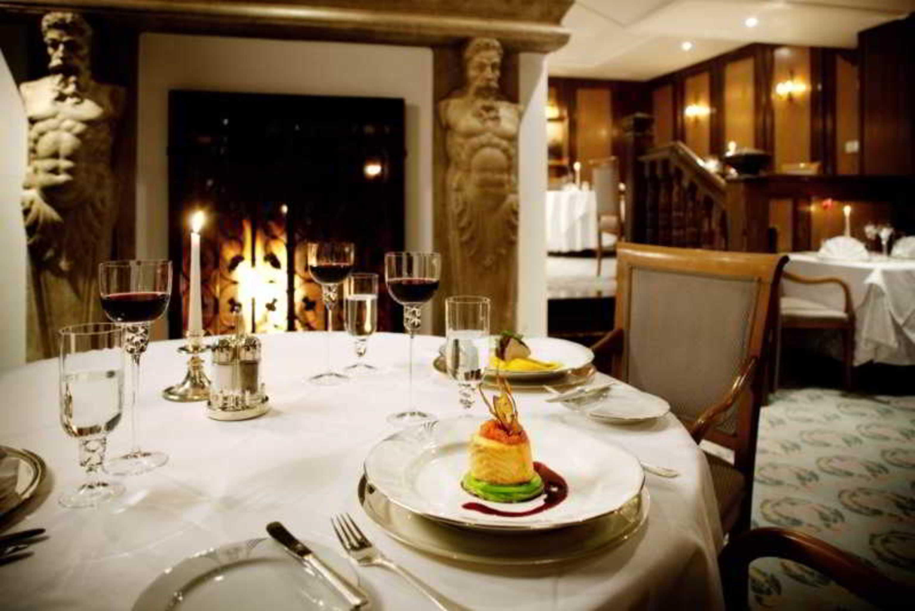 Art Nouveau Palace Hotel Praga Ristorante foto The photo depicts an elegant dining setting in a restaurant. In the foreground, there is a round table elegantly laid with fine dining utensils, including plates, cutlery, and glasses. 

On the table, a beautifully presented dessert sits on a plate, 