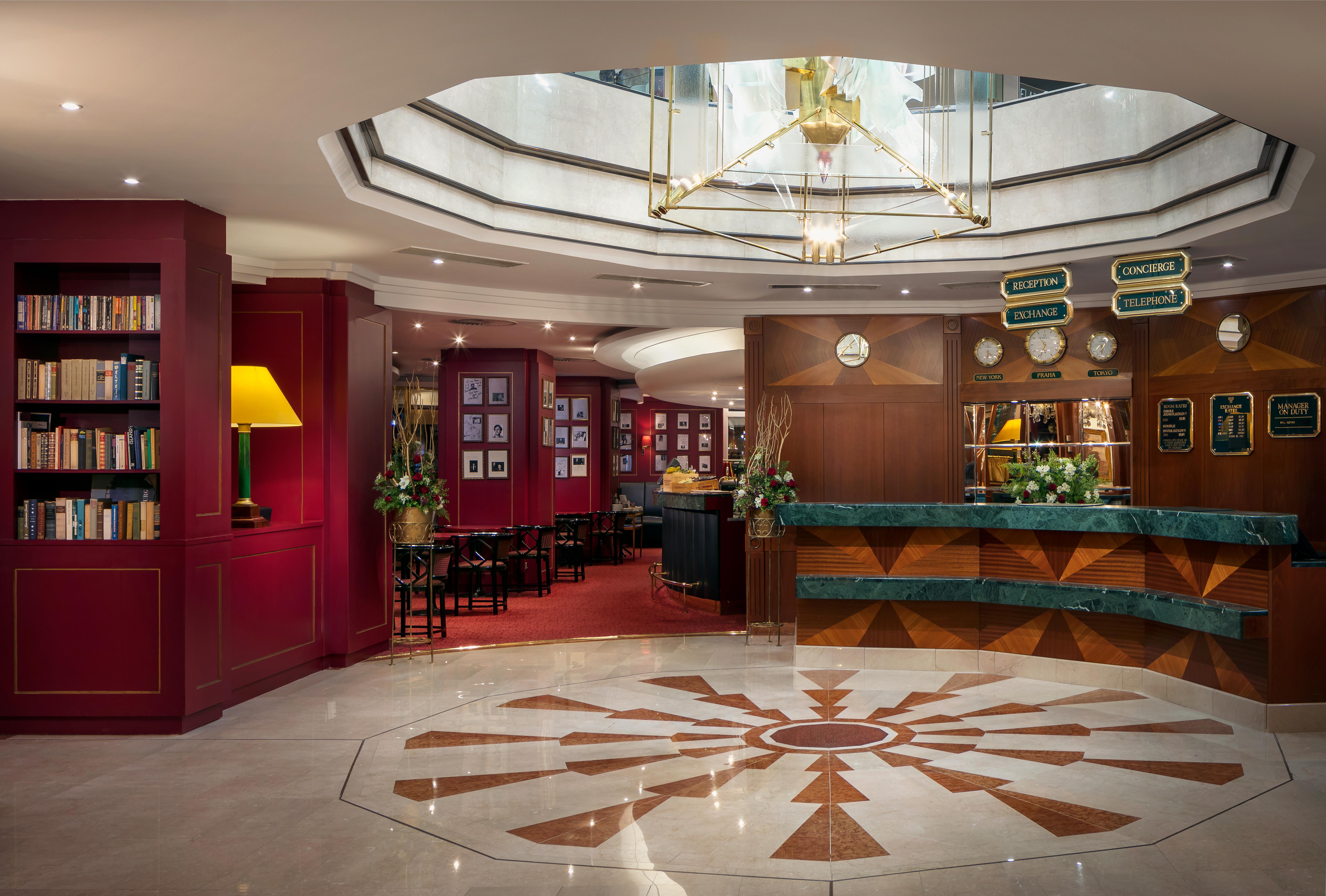 Art Nouveau Palace Hotel Praga Esterno foto The image shows the interior of a hotel lobby. The space features a stylish and elegant design with rich red walls and a polished marble floor that has a decorative pattern. There's a reception desk with a green marble top and a warm, welcoming area 