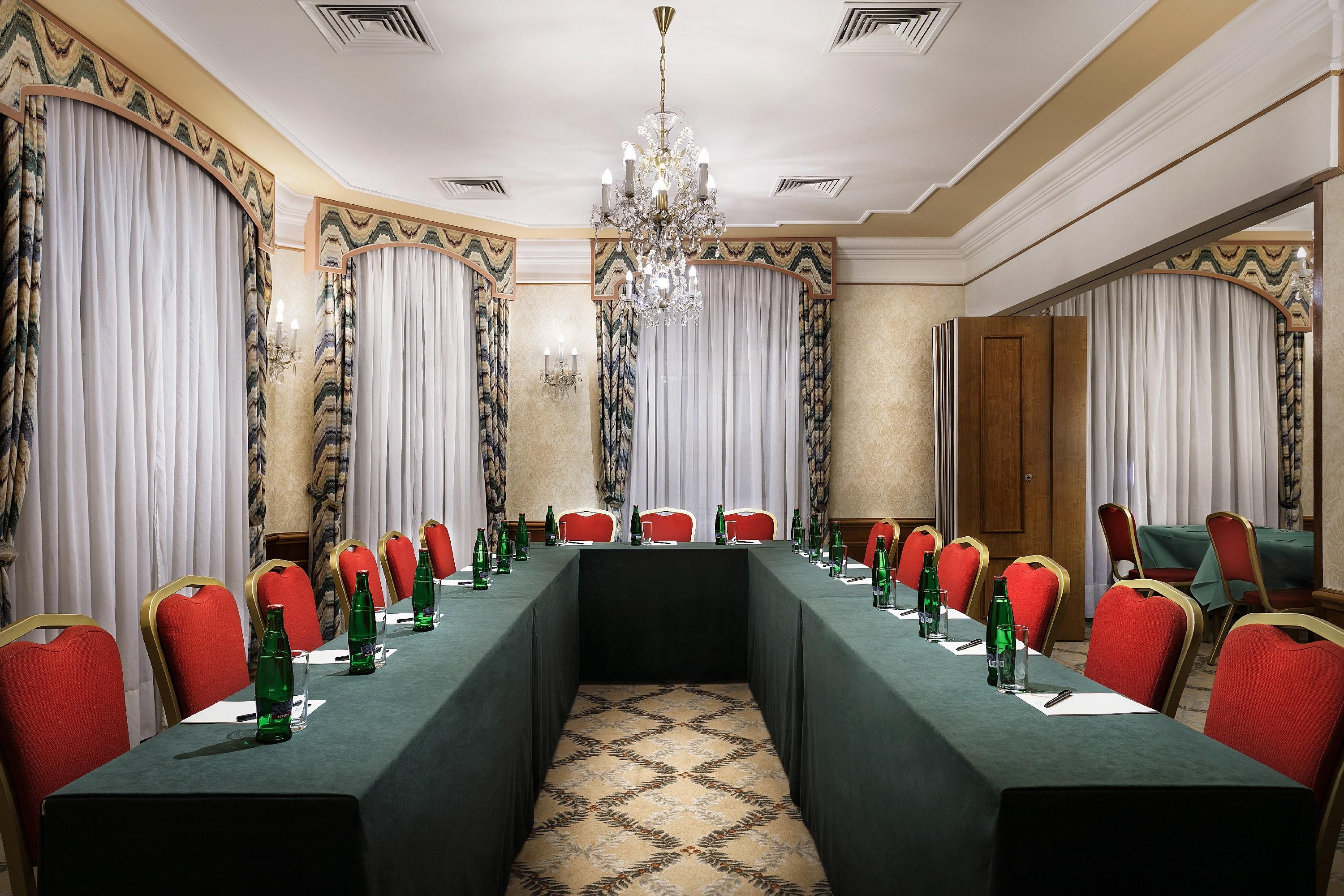 Art Nouveau Palace Hotel Praga Esterno foto The photo shows a conference room set up for a meeting. There is a rectangular arrangement of tables covered with green cloth, surrounded by red chairs. Each place has a notebook and an unfinished glass bottle of water. The room features elegant deco