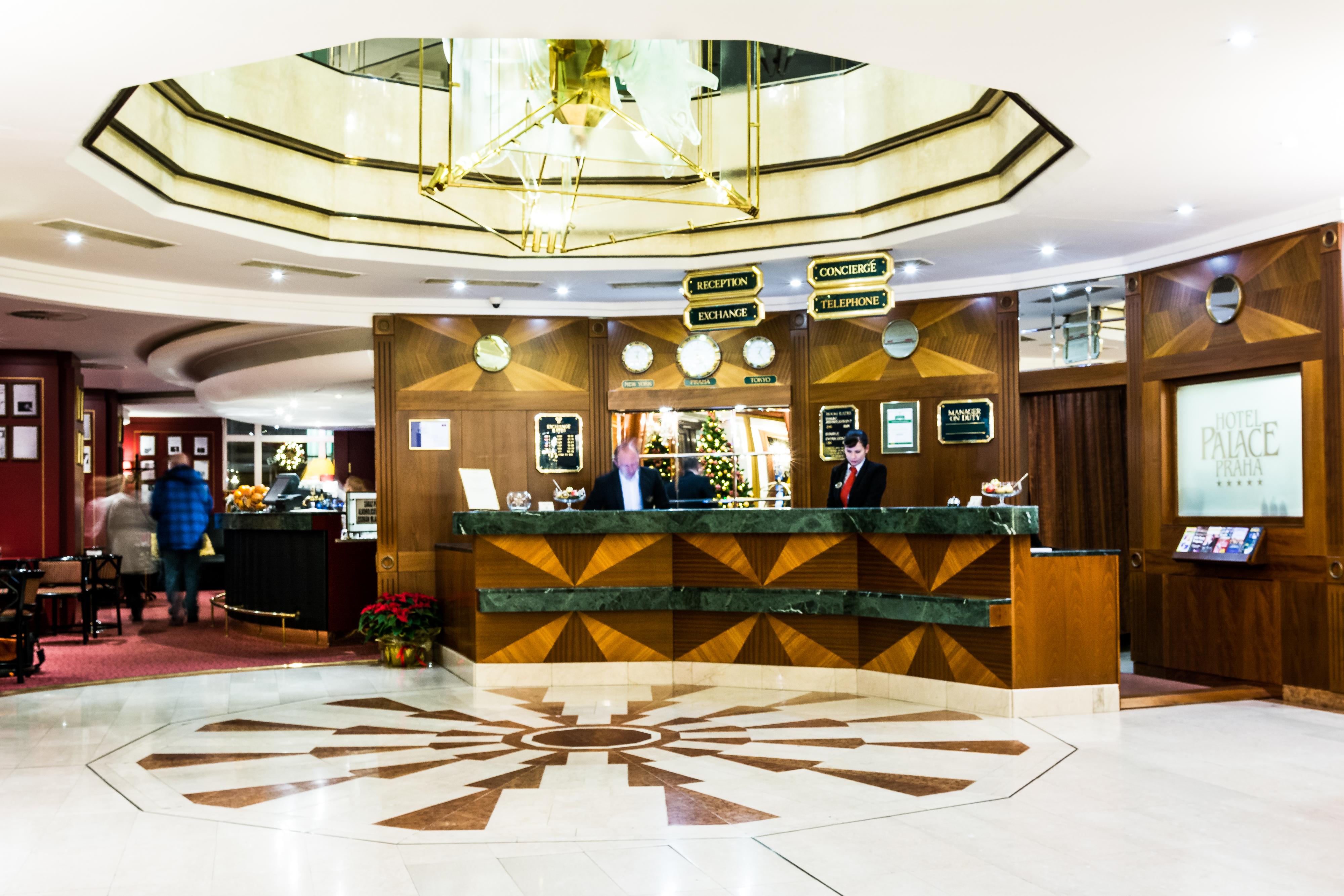 Art Nouveau Palace Hotel Praga Esterno foto The photo depicts the interior of a hotel lobby. At the center, there is a reception desk made of dark wood with a decorative design. Behind the desk, two staff members are standing, likely ready to assist guests. Above the reception area, there are 