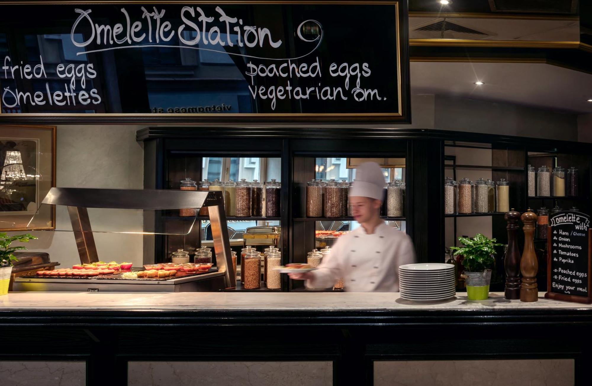 Art Nouveau Palace Hotel Praga Esterno foto The photo shows a restaurant or hotel breakfast area, specifically an "Omelette Station." There is a chalkboard sign that lists menu items such as "fried eggs," "omelettes," "poached eggs," and "vegetarian options." In the foreground, a chef wearing 