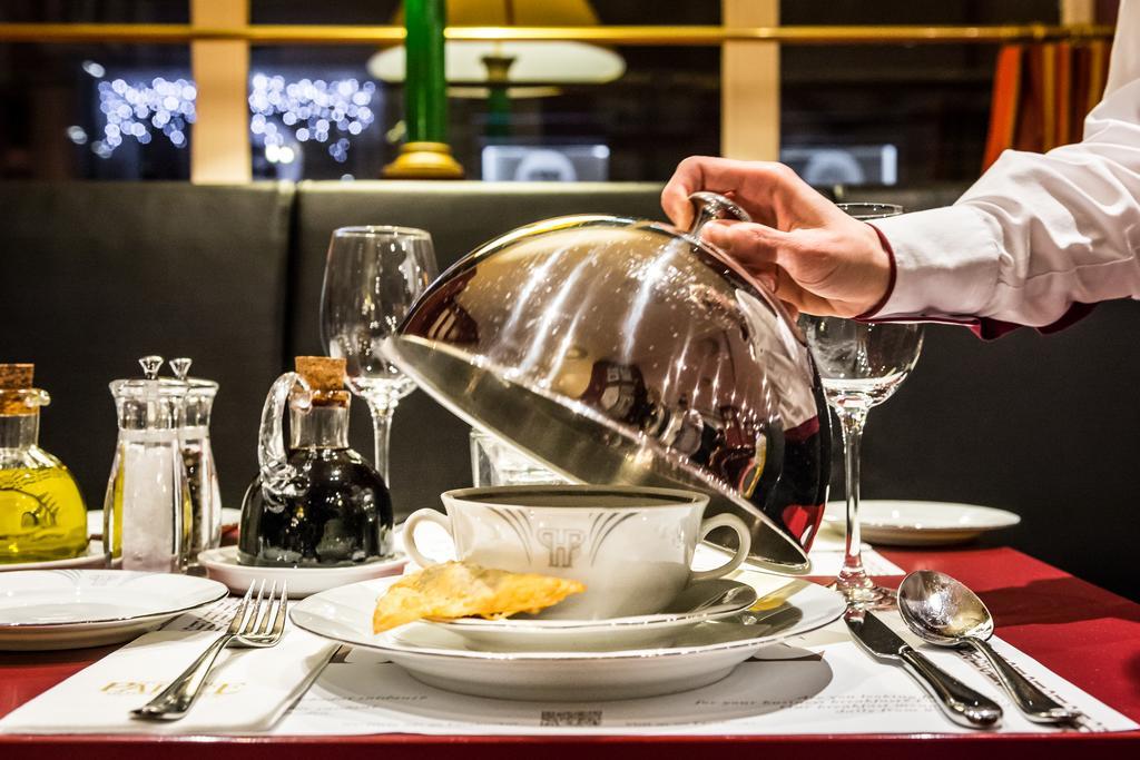 Art Nouveau Palace Hotel Praga Esterno foto The photo shows a beautifully set dining table with a focus on a server lifting a cloche away from a bowl of food. The bowl is placed on a decorative plate, and there is a crispy item resting on the plate. Surrounding the bowl are elegant dining uten