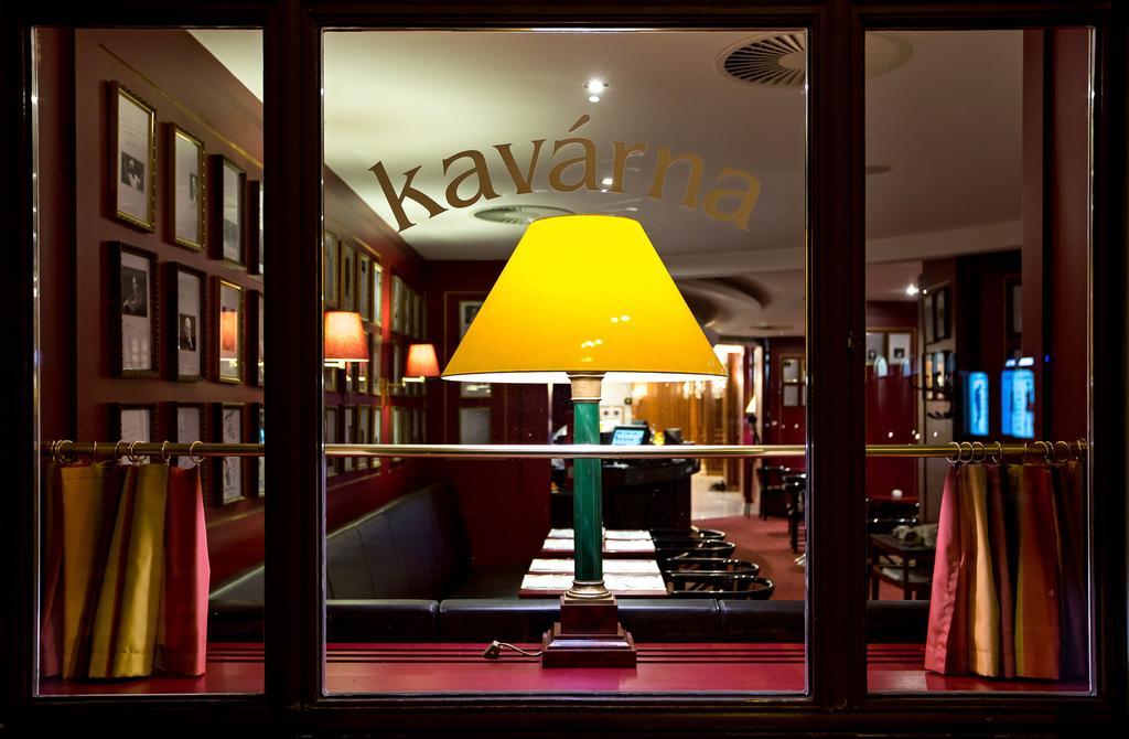 Art Nouveau Palace Hotel Praga Esterno foto The photo appears to show the interior of a café or restaurant through a large window. The word "kavárna," which means "café" in Czech, is visible on the window. Inside, there is a cozy atmosphere with a distinct yellow lamp glowing on a table, provi