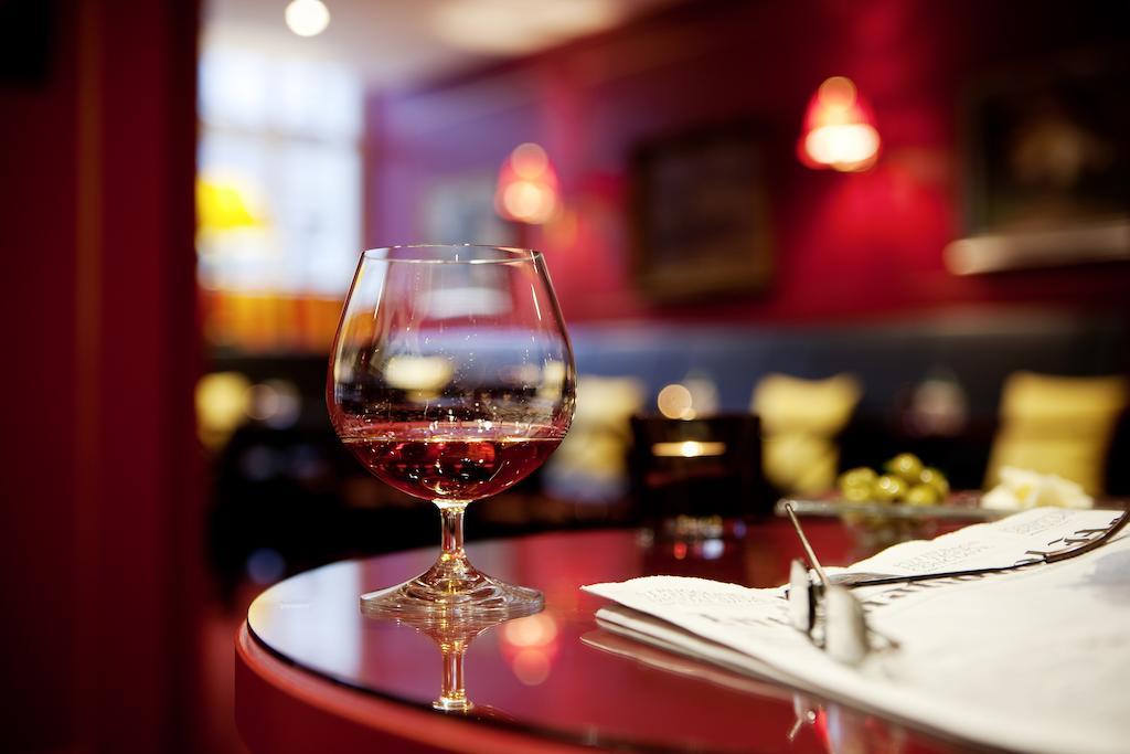 Art Nouveau Palace Hotel Praga Esterno foto The photo shows a sophisticated scene in a cozy bar or restaurant. In the foreground, there is a glass of red wine resting on a polished table. The glass has a round bowl and sits on a short stem. In the background, there are soft, warm lighting fixt