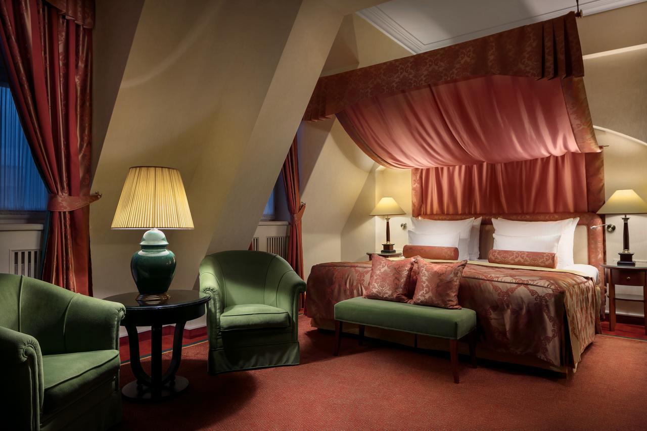 Art Nouveau Palace Hotel Praga Esterno foto The photo depicts a cozy and elegantly furnished hotel room. It features a bed with a canopy draped in pink fabric, complemented by rich red and gold decor. There are two green armchairs and a small round table near the bed, creating a relaxed seatin